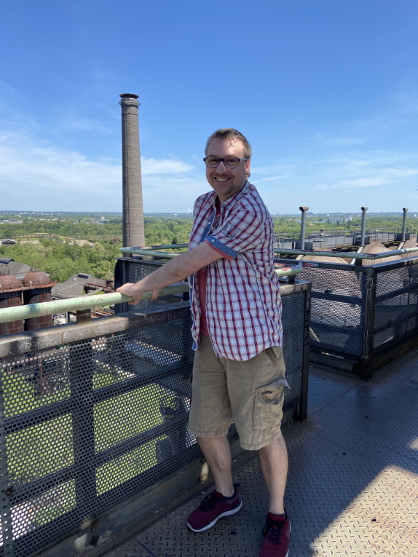 André auf dem Industriedenkmal eines Hochofens. Er hält sich krampfhaft an einem Geländer fest. Im Hintergrund überwucherte Industrieanlagen und ein Schornstein.