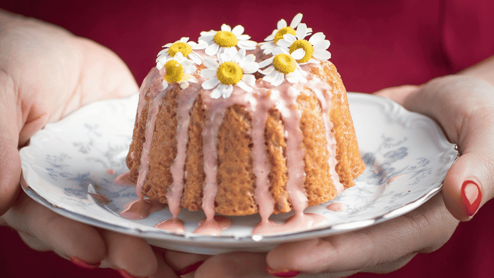 Frauenhände halten einen Teller, auf dem sich ein mit Blüten verzierter kleiner Kuchen befindet