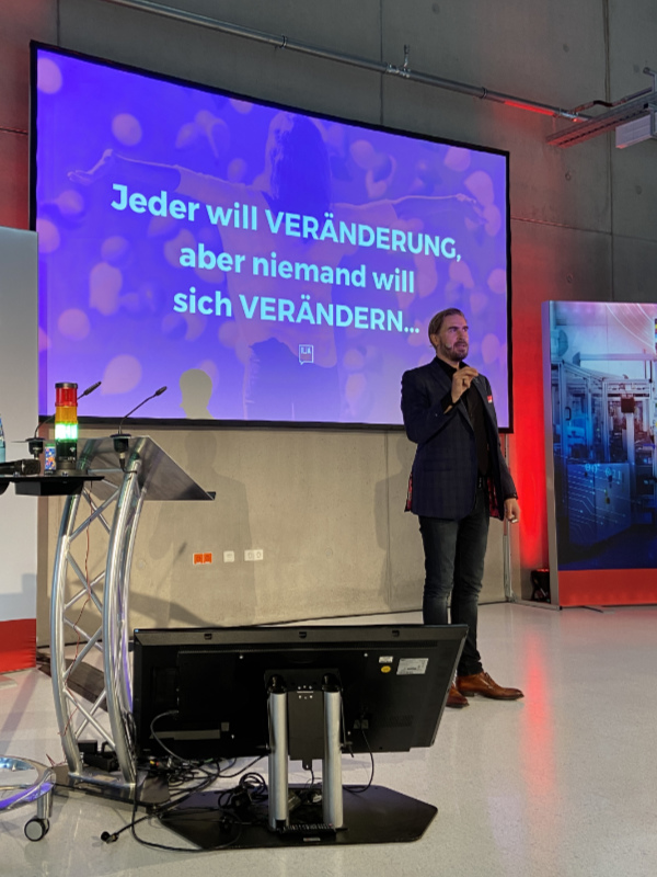 Vortragsredner Ilja bei einer Keynote vor einer Leinwand, auf der steht: "Jeder will Veränderung, aber niemand will sich verändern..."