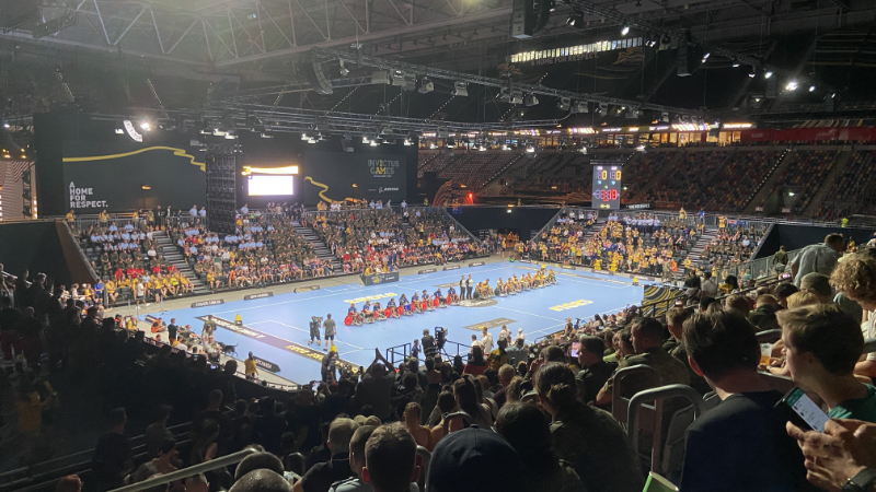 Blick von der Tribüne auf das Spielfeld eines Rollstuhlrugby-Spiels. Die Spieler sind für die Nationalhymne aufgereiht in der Mitte des Spielfeldes.